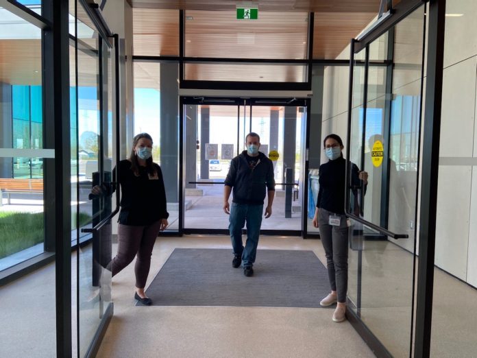 Deux femmes qui ouvrent les portes d'un édifice vitré et un homme qui entre. Tous masqués.