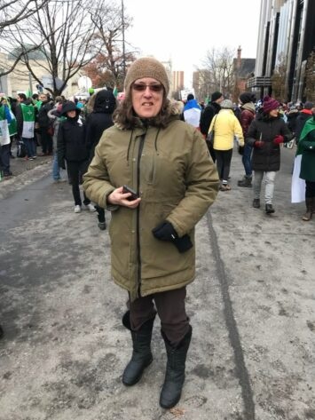 Une femme, vêtue d'un manteau, est debout devant une foule dans les rues d'Ottawa. Certaines personnes dans la foule portent des drapeaux franco-ontariens sur leurs épaules.