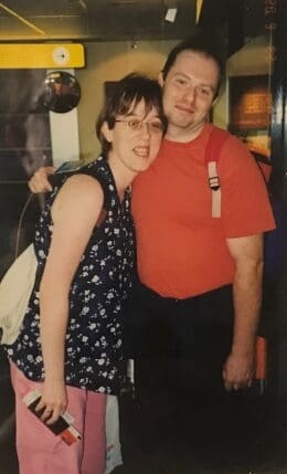 Une femme et un homme portant des vêtements d'été, s'enlacent en souriant au photographe. Ils portent de légers sacs à dos et la femme tient un passeport et un billet d'avion dans sa main