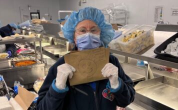 Femme portant un masque, un filet de protection sur la tête et des gants dans un espace de préparation des repas, tenant dans ses mains le dessin d'un bonhomme-sourire