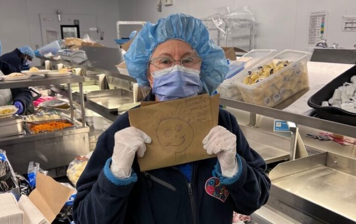 Femme portant un masque, un filet de protection sur la tête et des gants dans un espace de préparation des repas, tenant dans ses mains le dessin d'un bonhomme-sourire