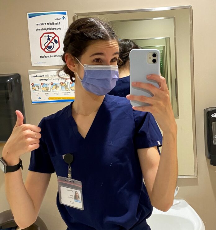 Jeune femme masquée et portant des "scrubs" qui prend un selfie dans une salle de bain de Montfort avant son premier quart de travail